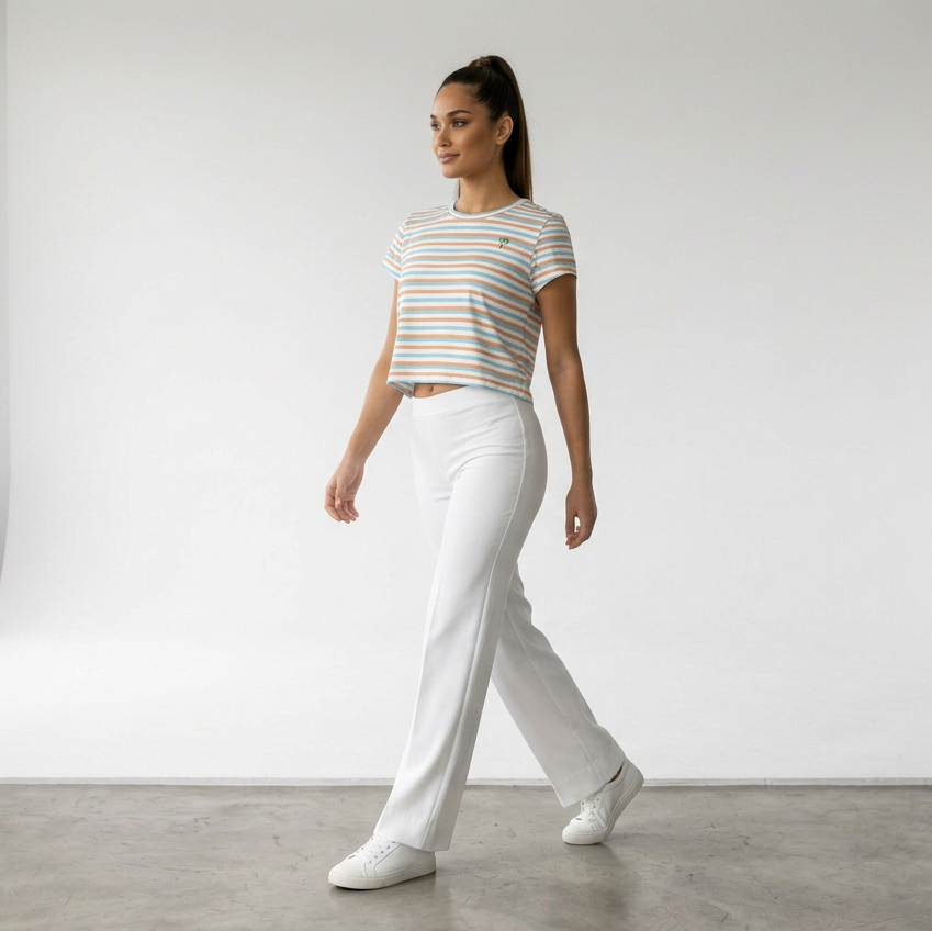 A woman walking in a bright white studio wearing a short-sleeve striped crop top in white, orange, and light blue, paired with white pants and white sneakers.