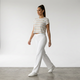 A woman walking in a bright white studio wearing a short-sleeve striped crop top in white, orange, and light blue, paired with white pants and white sneakers.