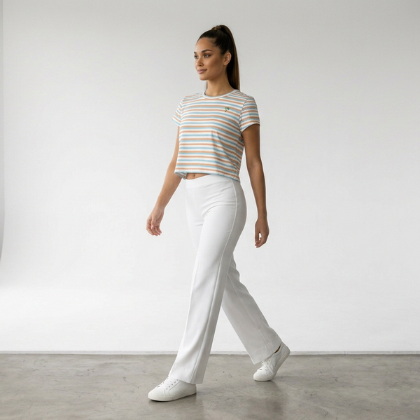 A woman walking in a bright white studio wearing a short-sleeve striped crop top in white, orange, and light blue, paired with white pants and white sneakers.